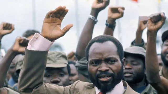 Samora Machel é um homembet nacional regraspele escura, cabelos bem rentes. Estábet nacional regrasmeio a vários homens, todos com o braço direito erguido.