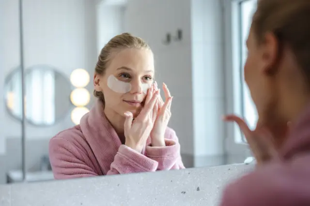 Kobieta patrzy w lustro, nakładając płatki pod oczy. Ma na sobie różowy szlafrok.