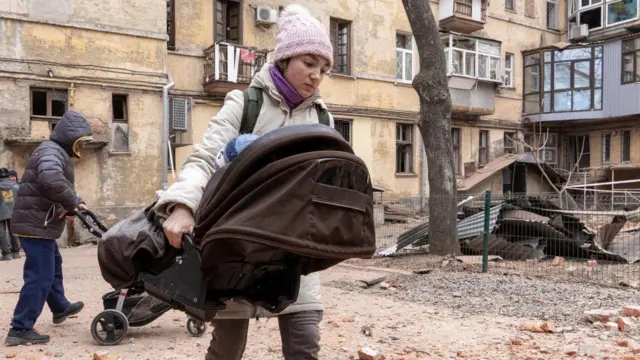 Матір із дитиною після обстрілу в Харкові, 2023 рік. Ззаду з коляскою чоловік. Вони в дворі понівеченого будинку