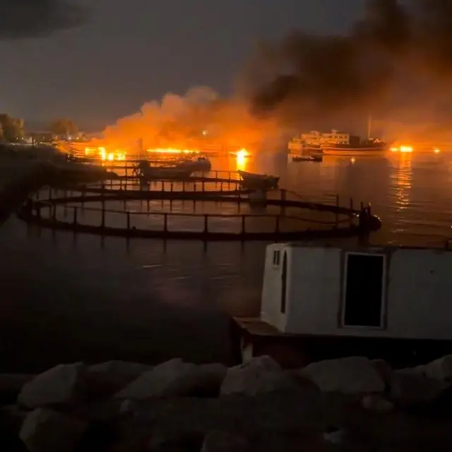 Liman boyunca uzanan kıyı şeridinde çıkan yangının gece görüntüsü. Parlak turuncu alevler ve yoğun siyah duman, kıyı şeridinin çeşitli noktalarından yükseliyor ve su yüzeyine yansıyor. Yangının yakınında birkaç tekne görülebiliyor, ön planda ise dairesel deniz yapıları ve kayalar yer alıyor.