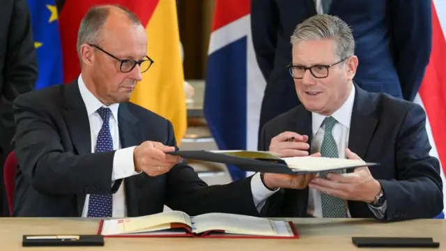 ダークスーツにめがね姿の2人の男性がテーブルに座り、署名した条約のファイルを交換している。テーブルの後ろにはドイツとEU、イギリスの旗が立てられており政府職員が立っている