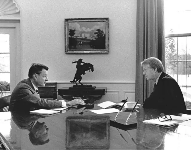 برژینسکی و کارتر ۱۹۷۷ در کاخ سفید مقابل هم نشستهاند و صحبت میکنند تصویر آنها روی میز منعکس شده