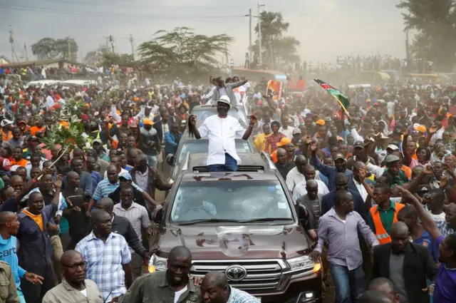 Raila Odinga sits on the roof of his car, surrounded by supporters.