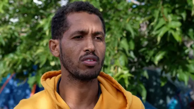 A man in a light orange hoodie talks during an interview