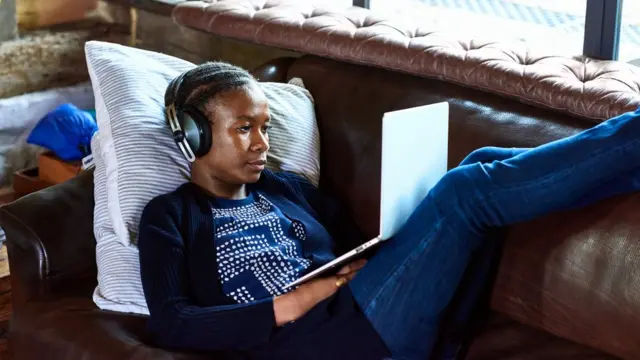 Mulher usando laptop e fonescasa de aposta nordeste futebolouvido enquanto está deitadacasa de aposta nordeste futebolum sofá