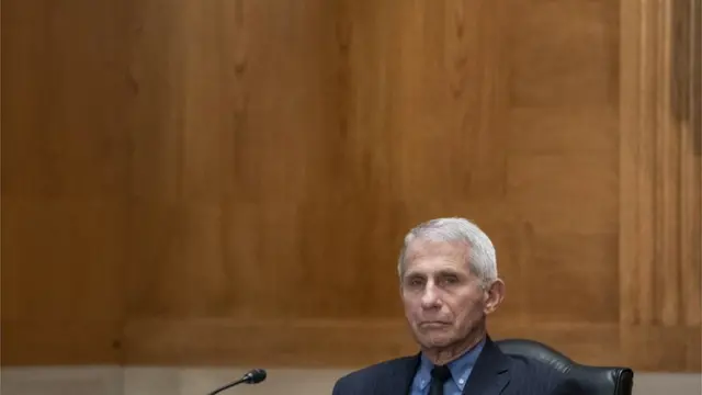 Anthony Fauci com feição séria sentado em frente a microfone, em sala de reunião