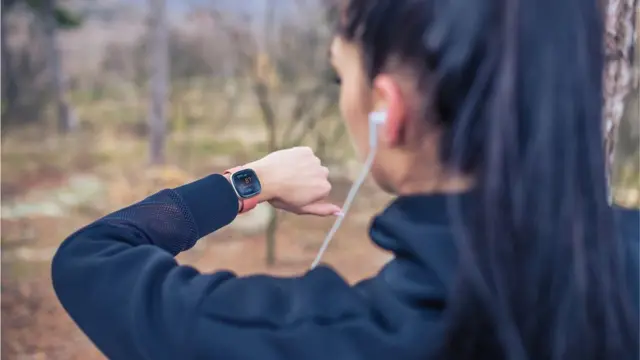 Fotografia colorida mostra mulhero jogo da roletinhapele branca e cabelo pretoo jogo da roletinhacostas, olhando um relógio do tipo smartwatch e usando roupas esportivas