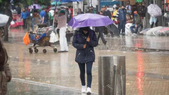 Moça de máscara e casaco de frio caminha sob a chuva com guarda-chuva, ao fundo, diversas pessoas na rua