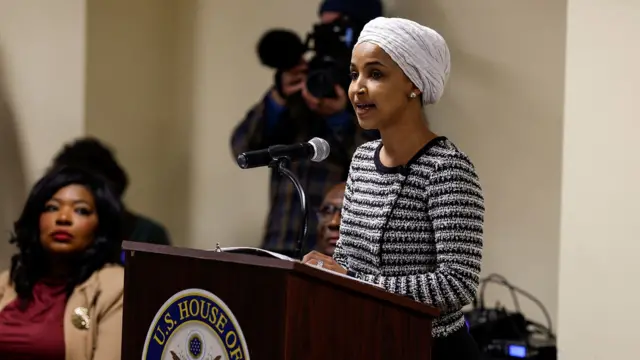 A photo of Olhan Omar addressing a town hall meeting while speaking into a microphone at a podium with others in the background, the podium displays a logo saying 'US House of'.