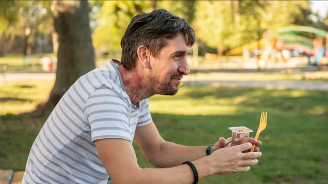 Muškarac u svetloj prugastoj majici sedi u parku i drži jogurt i viljušku u ruci