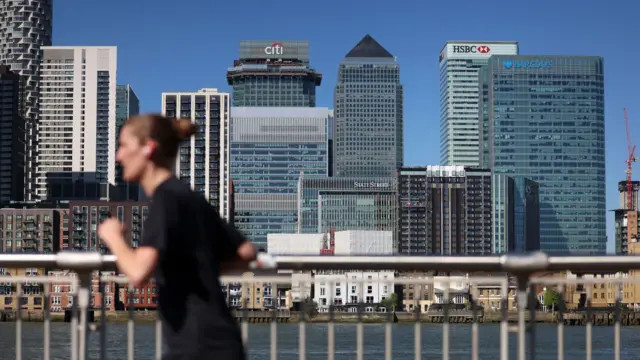 倫敦金絲雀碼頭一位女士跑步而過，背景是銀行總部區天際線，可見匯豐銀行、巴克萊銀行、花旗銀行等的辦公大樓（7/4/2025）
