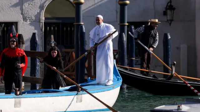 Muškarac obučen kao papa stoji na čamcu i u jednoj ruci drži veslo