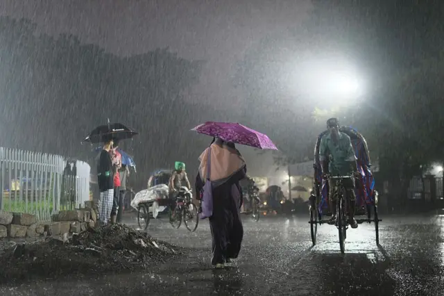 বৃষ্টির মধ্যে ছাতা মাথায় হেঁটে যাচ্ছেন বোরকা পরা এক নারী, রাস্তার একপাশে ছাতা মাথায় দাঁড়িয়ে আছেন কয়েকজন। কয়েকটি রিকশা যাচ্ছে রাস্তা দিয়ে