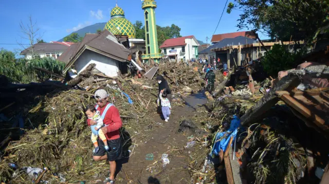 Warga berjalan di atas tumpukan material akibat banjir bandang di Jorong Galuang, Nagari Sungai Pua, Agam, Sumatera Barat, Senin (13/05/2024). 