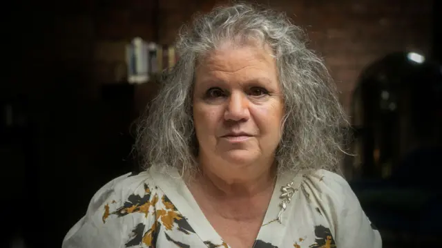 Ann Pearston — kobieta o długich, siwych włosach, ubrana w białą bluzkę w zielono-żółte kwiaty — pozuje w pomieszczeniu z ceglanymi ścianami. 