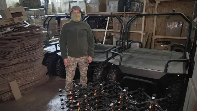 Mężczyzna stoi w magazynie obok ułożonych w stos dronów powietrznych i trzech dronów lądowych. Ma na sobie mundur wojskowy i twarz zasłoniętą zieloną tkaniną.