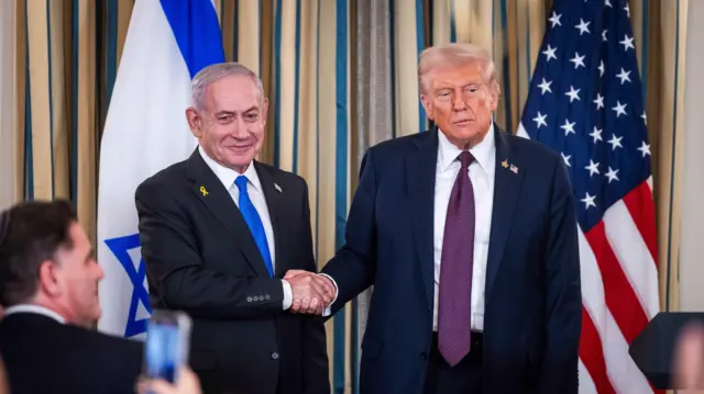 O primeiro-ministro de Israel, Benjamin Netanyahu, com sorriso no rosto, cumprimenta com aperto de mão o presidente dos EUA, Donald Trump, com um olhar sério em frente a repórteres na Casa Branca, após apresentação de plano de paz para Gaza. Atrás deles há bandeiras dos dois países.