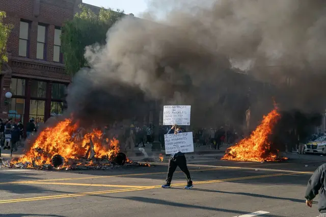 8 Haziran 2025 Pazar günü, ABD'nin Kaliforniya eyaletinin Los Angeles kentinde protestocuların arkasında arabalar yanıyor. Trump'ın hafta sonu Ulusal Muhafız birliklerini görevlendirmesi, yerel liderler ile yönetim arasında gerginliğin artmasına neden oldu.