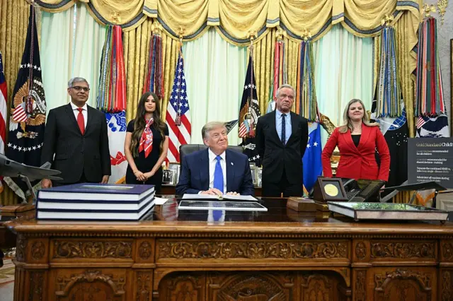 ডোনাল্ড ট্রাম্প বসে আছেন, পেছনে দাঁড়িয়ে আছেন কয়েকজন