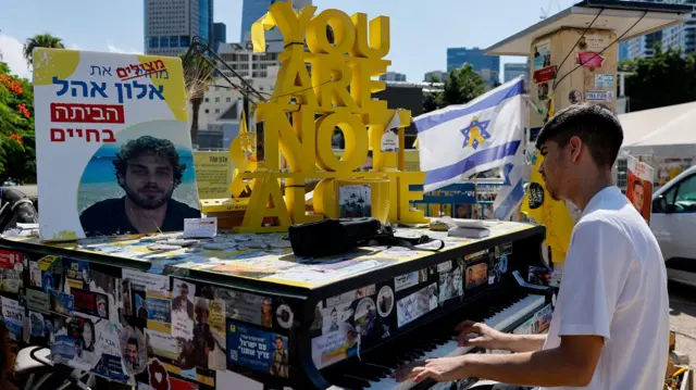 Акция с требованием освободить заложников в Израиле: мужчина играет на фортепиано, которое установлено на одной из площадей в Израиле. На инструменте множество наклеек с портретами заложников, на нем стоит плакат с фотографией заложника и желтая надпись "вы не одни"