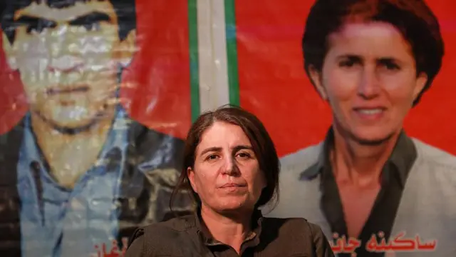 Kadın pkklı arkada iki fotoğrafın önünde kameraya bakıyor. oturmuş durumda