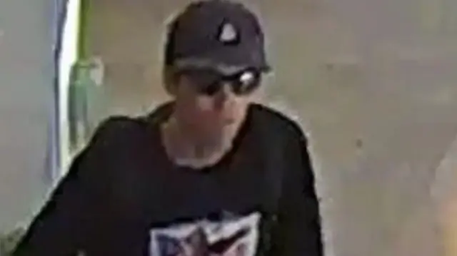 Un homme portant une casquette de baseball, des lunettes de soleil et une chemise bleue avec un motif. L'image est de faible résolution.