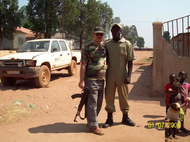  Двое мужчин в военной форме в Африке, один из них, в темных очках и европейского облика, держит в руке автомат