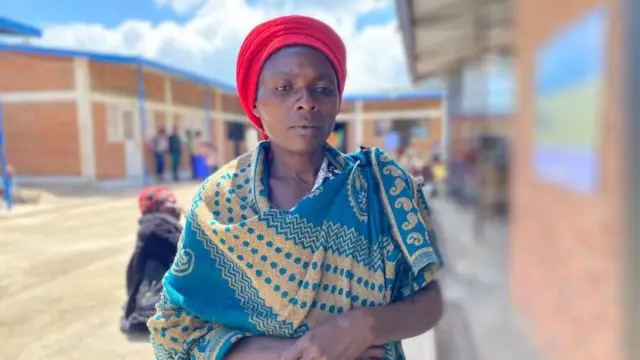 Umugore witeye igitenge ateze igitambaro gitukura, ahagaze yegeranyije ibiganza. 