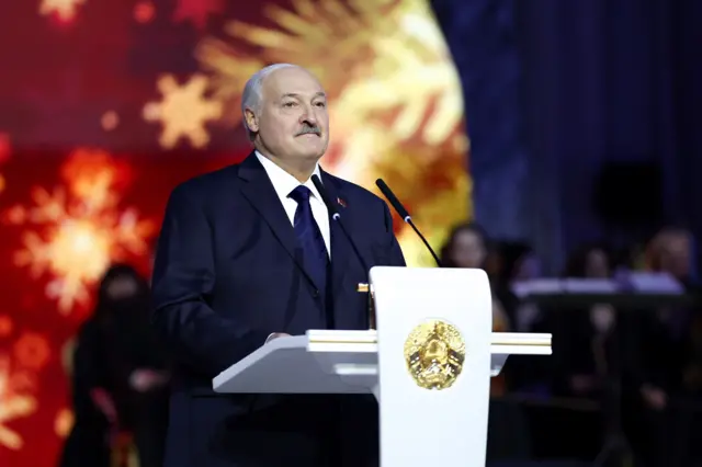 Александр Лукашенко в ходе одного из торжественных мероприятий в начале 2026 года. Он стоит в темно-синем костюме на сине-красно-золотом фоне, перед белой трибуной с позолоченным гербом Беларуси на ней