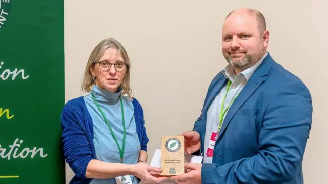 Clare Topping is presented with the award by Stuart Dawks. Ms Topping is wearing a blue polo-neck and blue cardigan and has a green lanyard around her neck. She is wearing glasses and has short blonde hair. Mr Dawks is wearing a blue jacket over a blue-striped shirt with a green lanyard around his neck. Between them they are holding a wooden award. They are both smiling at the camera.
