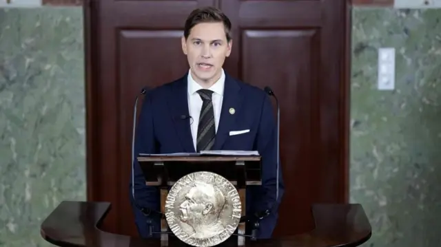 ノーベル賞の創設者アルフレッド・ノーベルの横顔が刻まれたプレートのついた演台に向かう、フリドネス委員長。紺のスーツに斜めしまのネクタイを締めている