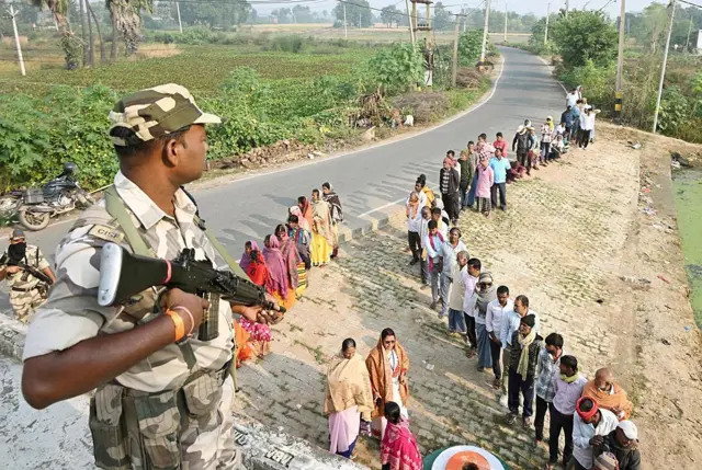 একটি রাস্তার পাশে নারী ও পুরুষ ভোটারদের দুটি লাইন। নারীদের লাইনের পাশে একজন এবং ছাদের ওপর আরেকজন নিরাপত্তারক্ষী অস্ত্র হাতে দাঁড়িয়ে আছেন