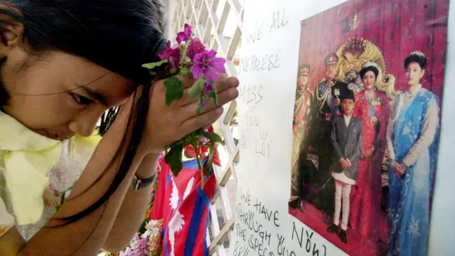 Młoda dziewczyna w trzyma fioletowe kwiaty i modli się przed portretem zmarłej nepalskiej rodziny królewskiej. Wokół zdjęcia na białym papierze widnieją napisy.