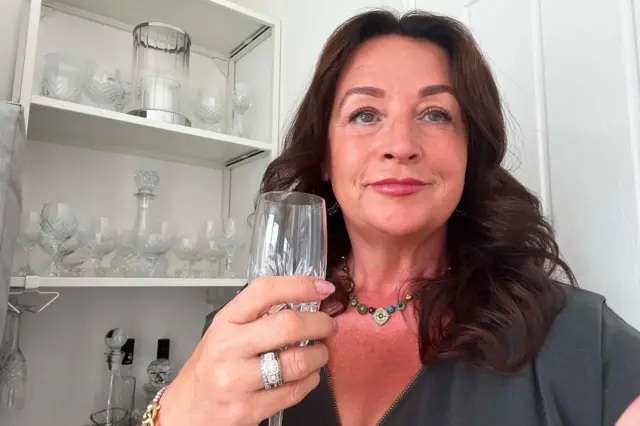 Mulher com longo cabelo escuro e expressão pensativa, segura uma taça de champanhe, com um grande anel visível no dedo anular da mão que segura a taça