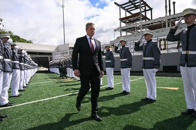 Дрісколл у темному діловому костюмі йде по зеленому штучному газону між двома шеренгами військових курсантів у сірій формі з білими брюками та кашкетами. Курсанти віддають честь. На задньому плані видно трибуни стадіону.