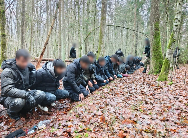 Задержанные мигранты сидят на корточках в лесу длинной шеренгой. У них за спиной ходят польские пограничники