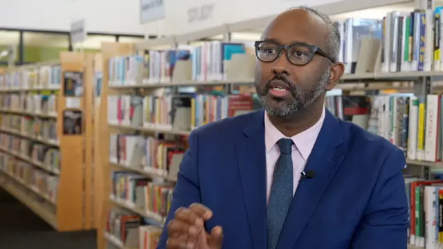 Une photo de Jaylani Hussein en costume bleu, cravate bleue et lunettes, gesticulant face à la caméra dans une bibliothèque dont on aperçoit les étagères en arrière-plan.
