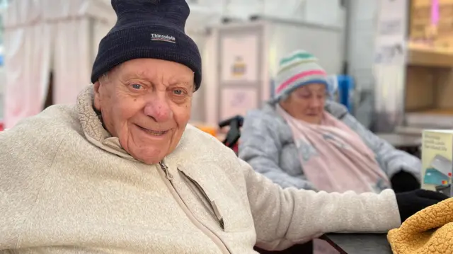 Dos personas están sentadas a una mesa en una zona con carpa y suelo rojo. Charles está en primer plano, con una chaqueta de lana color crema y un gorro de punto oscuro, recostado en una silla. Una manta amarillo mostaza cubre la mesa. Detrás de ellos, otra persona con un gorro de rayas de colores está sentada, y al fondo se ven puestos o casetas.
