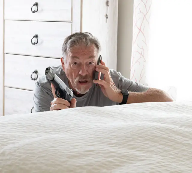 Un hombre sostiene un arma y un celular mientras hace una llamada escondido en una habitación