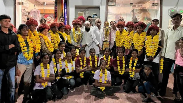 Uma equipe feminina de futebol aparece vestida com roupas esportivas e usando pompons amarelos. Dois homens que estão entre elas seguram troféus