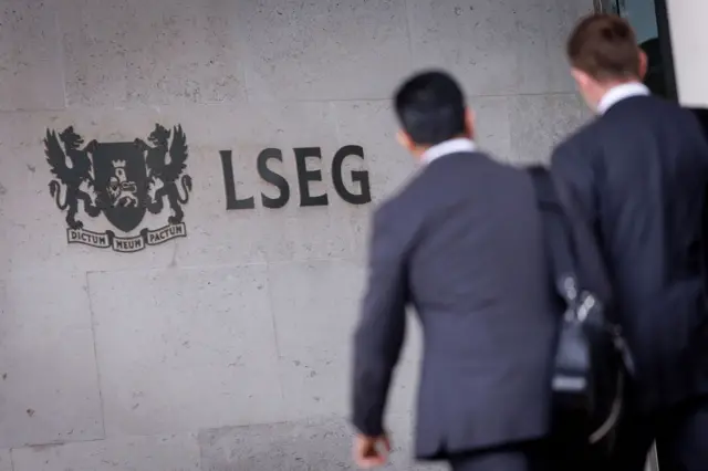 На заднем плане стена в серой плитке. На стене надпись LSEG. Рядом герб - два грифона в коронах держат щит. На щите изображен лев на фоне башни. Под щитом надпись на ленте dictum meum pactum. На переднем плане в сторону стены идут двое в костюмах.