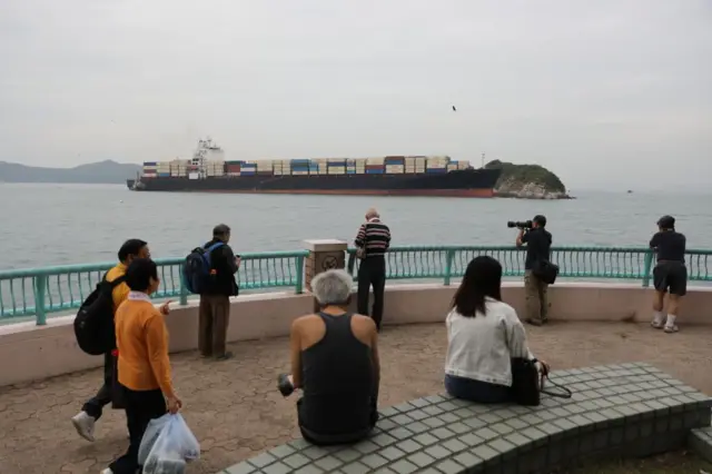 Un grupo de personas en un mirador hacia el mar. Frente a ellos, un gran barco mercante cargado de contenedores. 