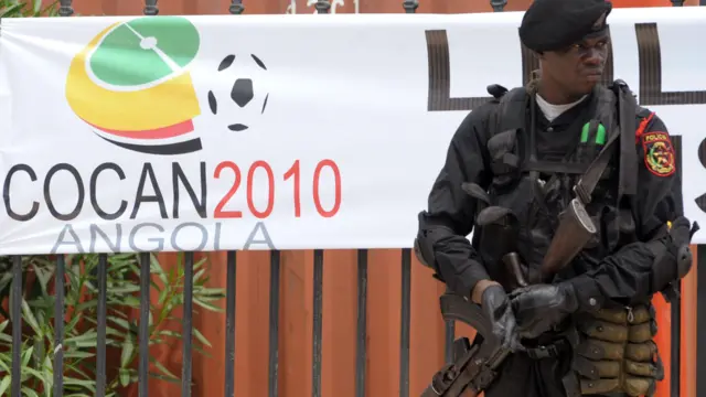 Un policier angolais, vêtu de noir avec un béret et un fusil automatique, monte la garde devant des grilles en fer qui portent une bannière avec le logo de la Coupe d'Afrique des Nations 2010.