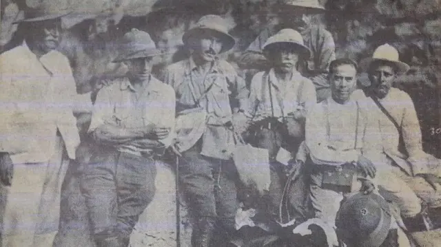 Siete hombres en trajes de exploradores y sombreros de safari posan frente a la cámara recostados sobre una roca