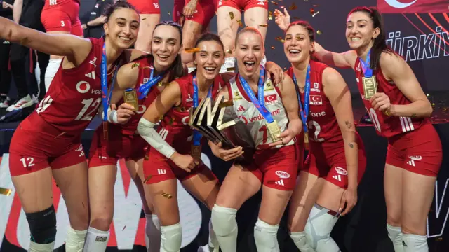 Voleybol milli takımı oyuncuları podyumda kupayı kaldırırken gülümsüyor, havada konfetiler uçuşuyor
