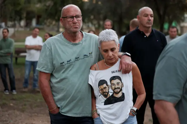 мужчина обнимает за плечи женщину, она в белой майке, на которой напечатаны фотографии двух молодых мужчин