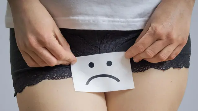 A close up of woman holding paper with sad face symbol  in front of her vagina