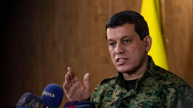 Mazlum Abdi askeri üniforması ve sarı bir bayrak önünde mikrofona konuşuyor