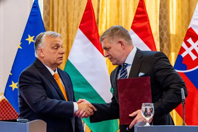 Орбан потискає руку Фіцо, який тримає у руці бордову папку. Вони стоять за трибунами на пресконференції на тлі прапорів Угорщини, Словаччини та ЄС
