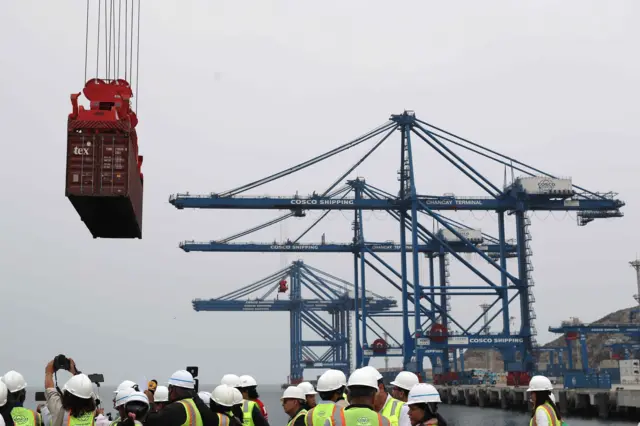 Grúas transportan un contenedor en el gran puerto de Chancay. Debajo, en el suelo, un grupo de personas con cascos y chalecos reflectantes. 